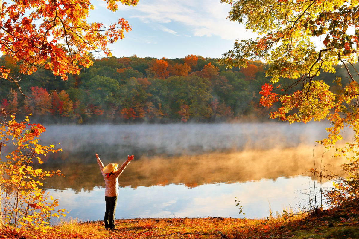 Person with arms raised at a misty lake framed by autumn foliage